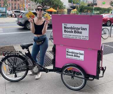person with black cargo bike with pink cargo boxes labeled "nonbinarian book bike"